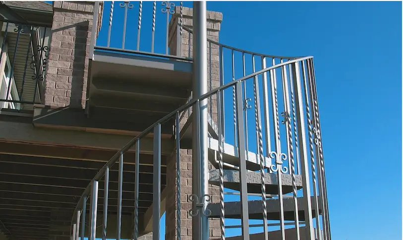 A spiral metal staircase with decorative railings, crafted by a skilled deck builder, is attached to a brick building and extends upward against a clear blue sky.