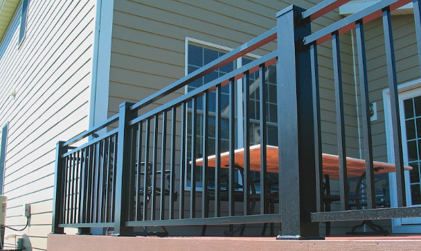 Black metal deck railing with vertical spindles surrounds a wooden patio deck. A table and chairs sit behind the railing, perfect for relaxing after a Deck Install. The house exterior features tan siding and large windows.