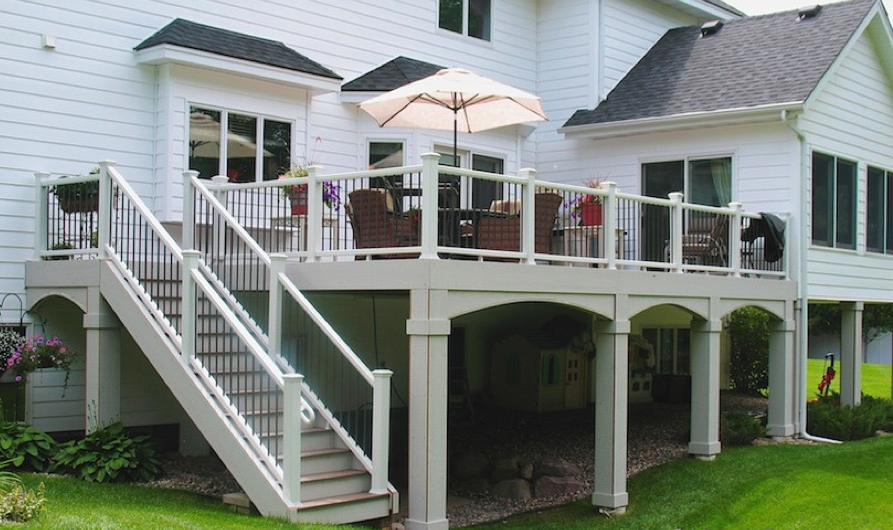 A raised white deck attached to a house features DIY deck materials, a stairway, railing, outdoor furniture, a table with an umbrella, and green lawn below. There’s a shaded area under the deck with kids’ play equipment.