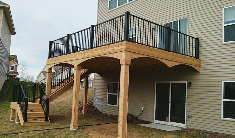 A two-story beige house with a wooden elevated deck, built with quality DIY Deck Materials and sleek black railings. The staircase leads to the yard, while the open area beneath creates a sheltered space above a patio door.