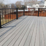 Spacious, empty backyard deck with light gray wood planks and black metal railings, perfect for DIY deck materials or a professional deck install, overlooking a grassy yard, trees, neighboring houses, and a wooden fence.