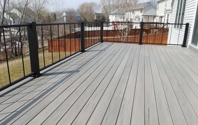 Spacious, empty backyard deck with light gray wood planks and black metal railings, perfect for DIY deck materials or a professional deck install, overlooking a grassy yard, trees, neighboring houses, and a wooden fence.