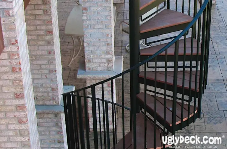 A black metal spiral staircase with wooden steps curves upward next to brick pillars on a stone patio, showcasing quality DIY Deck Materials. The logo uglyDeck.com appears in the lower right corner.
