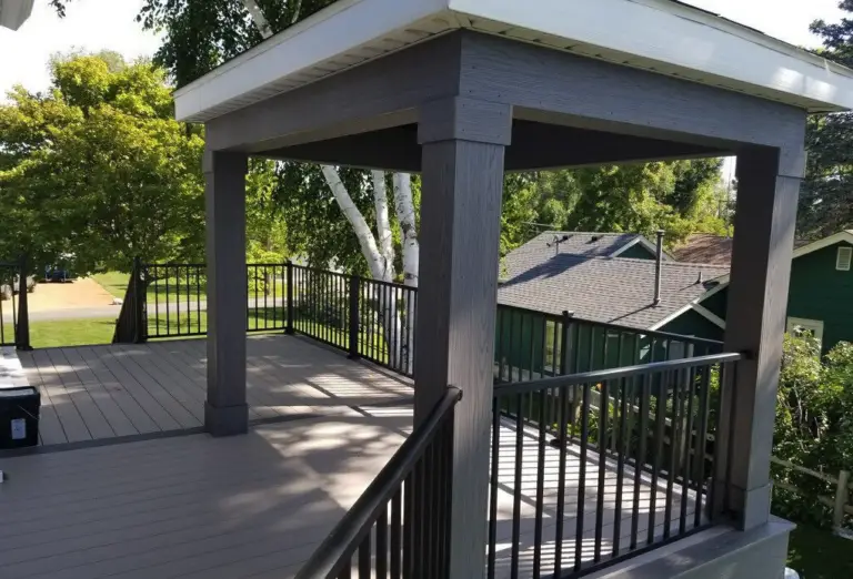 A covered outdoor deck with gray flooring, dark wooden posts, and black metal railings, built with quality DIY deck materials, overlooks a residential neighborhood surrounded by greenery and trees.