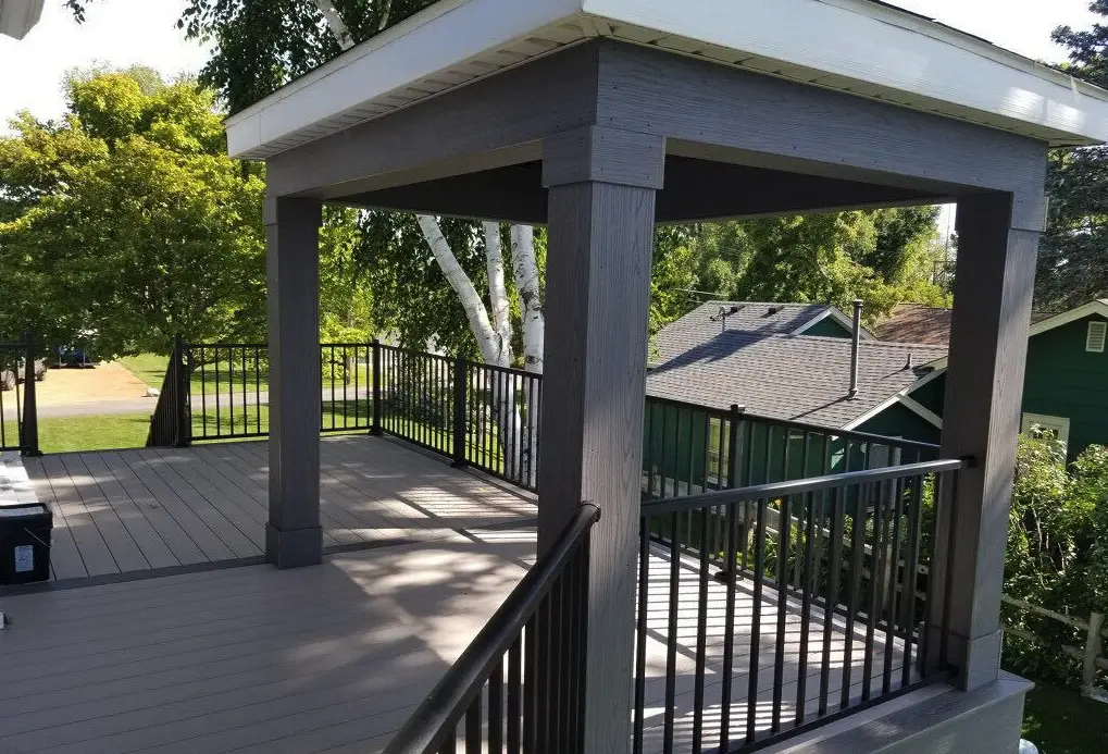 A covered outdoor deck with gray flooring, dark wooden posts, and black metal railings, built with quality DIY deck materials, overlooks a residential neighborhood surrounded by greenery and trees.