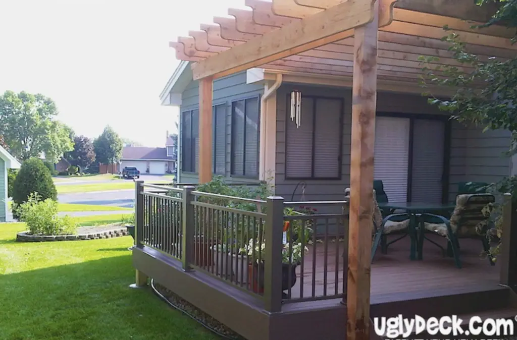 A backyard deck with a wooden pergola, metal railing, and patio furniture; expertly crafted by a Deck Builder, the deck is attached to a house and overlooks a green lawn and suburban neighborhood. uglydeck.com is visible in the bottom right corner.