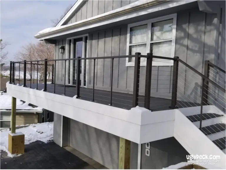 A modern gray house features a raised deck with dark railings and cable wires, all dusted with snow. Perfect for showcasing DIY Deck Materials or a Deck Install, the uglydeck.com logo appears in the corner.