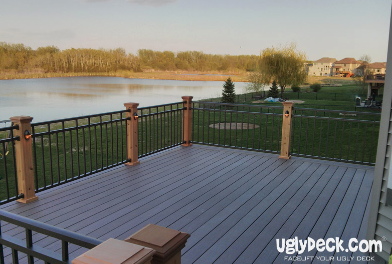 A spacious, modern deck built with quality DIY deck materials features wooden railings and overlooks a green lawn and tranquil lake, with houses and trees in the background. uglyDeck.com is displayed in the lower right corner.