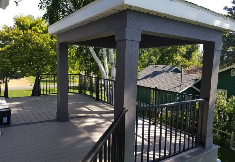 A wooden deck with dark railings and a covered pergola structure, crafted by a professional deck builder, is nestled outdoors among green trees and neighboring houses on a sunny day.
