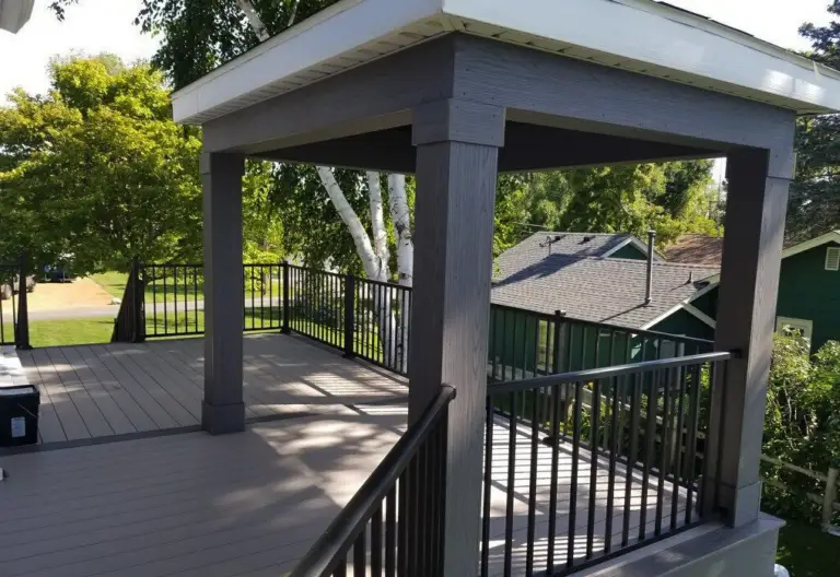 A wooden deck with dark railings and a covered pergola structure, crafted by a professional deck builder, is nestled outdoors among green trees and neighboring houses on a sunny day.