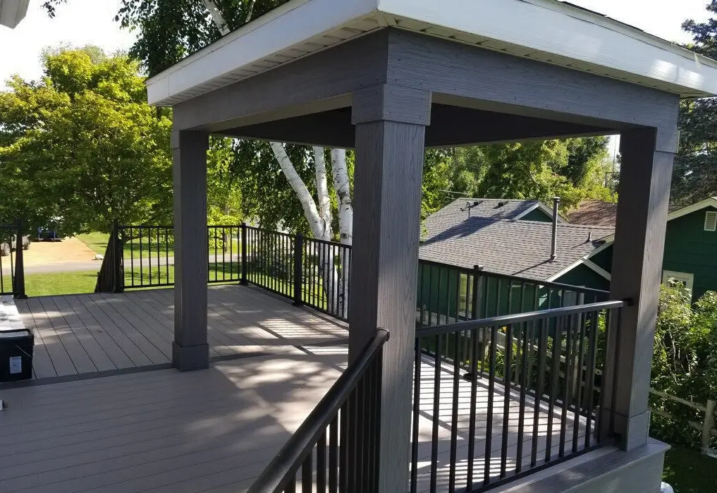 A wooden deck with dark railings and a covered pergola structure, crafted by a professional deck builder, is nestled outdoors among green trees and neighboring houses on a sunny day.
