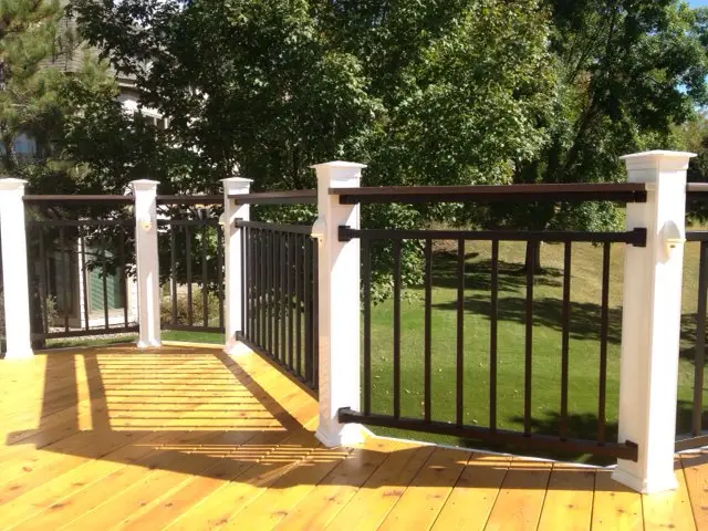 A wooden deck installed with white and black railings overlooks a green lawn and trees on a sunny day. Shadows from the railing are cast onto the deck’s surface, showcasing the expertise of a professional deck builder.
