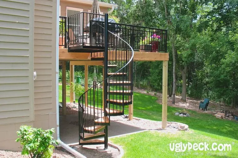 A black metal spiral staircase leads from a raised deck to a grassy backyard. The deck, built with quality DIY Deck Materials, has a grill, flowers, and black railing, and is attached to a beige house surrounded by lush green trees and lawn.