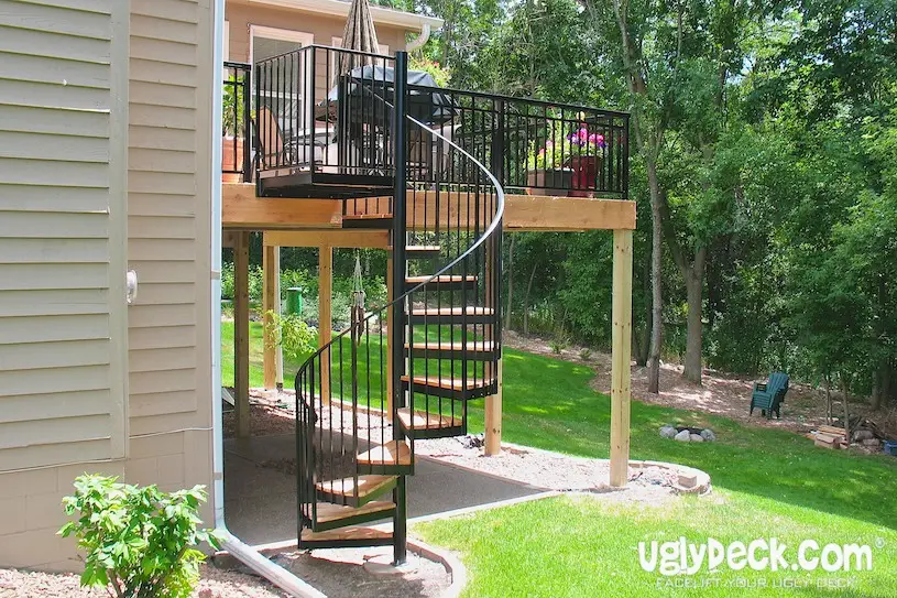 A black metal spiral staircase leads from a raised deck to a grassy backyard. The deck, built with quality DIY Deck Materials, has a grill, flowers, and black railing, and is attached to a beige house surrounded by lush green trees and lawn.