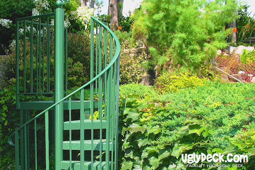 A green metal spiral staircase stands in a lush garden filled with dense greenery, leafy plants, and various shrubs—a perfect inspiration for your next DIY deck materials project. The text uglydeck.com appears in the bottom right corner.
