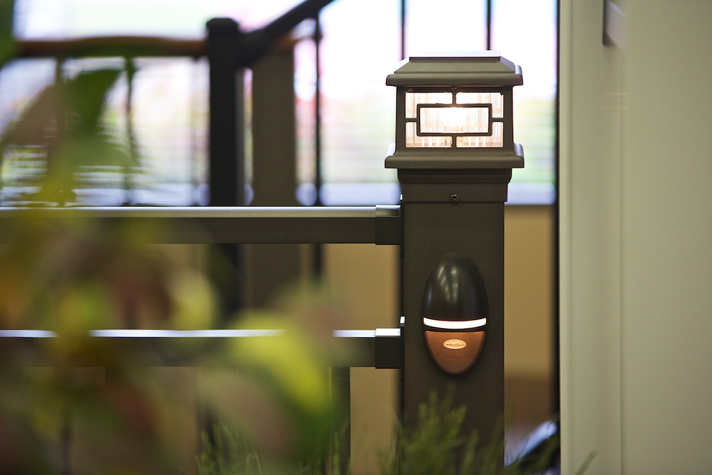 A close-up of a rectangular outdoor post light mounted on a dark metal railing, ideal for any DIY Deck Materials project. A small additional fixture sits below, with blurred plants and indoor railings in the background.