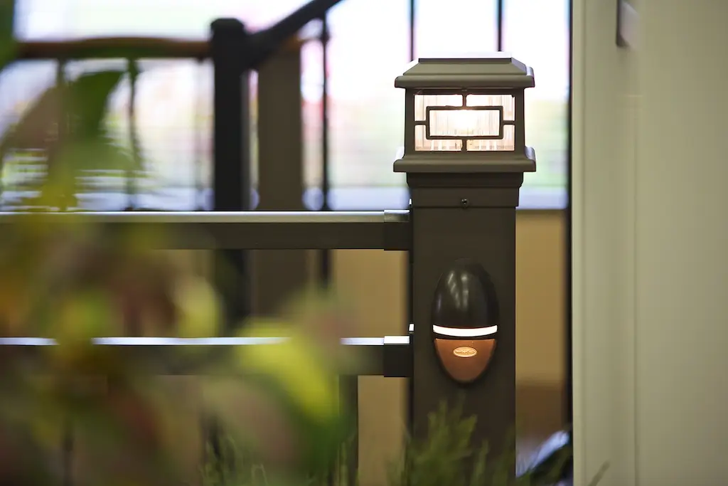 A close-up of a rectangular outdoor post light mounted on a dark metal railing, ideal for any DIY Deck Materials project. A small additional fixture sits below, with blurred plants and indoor railings in the background.