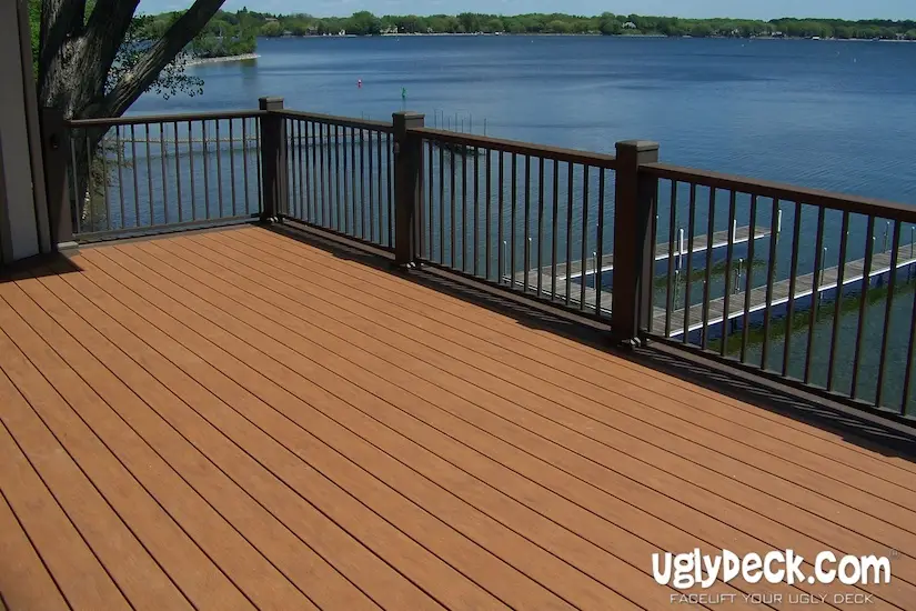 A spacious wooden deck with dark railings overlooks a calm lake, with docks and green trees visible across the water. Perfect for those seeking DIY deck materials or professional deck install services. The logo uglydeck.com is in the bottom right corner.
