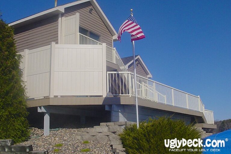 A tan house with a raised deck featuring white railings and privacy panels, built by expert Deck Builders. An American flag is mounted near the deck, and the yard has landscaping stones and green shrubs. UglyDeck.com is labeled in the corner.