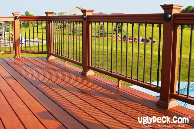 A wooden deck with metal railings overlooks a green lawn and landscaped backyard; the sunlit deck features uglydeck.com branding in the corner—perfect inspiration for your next DIY Deck Materials or professional Deck Install project.