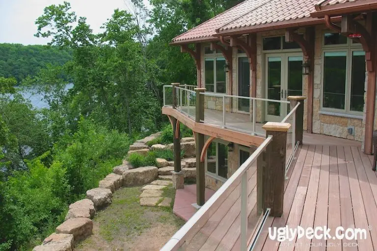 A wooden deck with glass railings wraps around a house overlooking a wooded area and river. Large windows line the house, and stone landscaping surrounds the deck. Deck Builder uglydeck.com is visible in the bottom right corner.