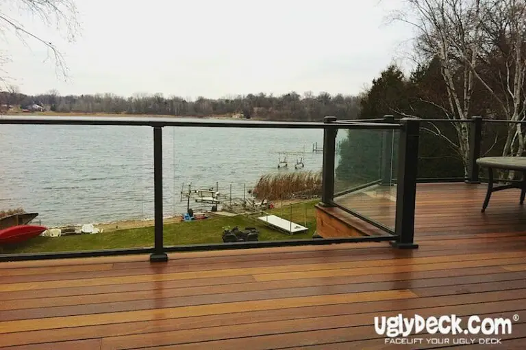 A wooden deck with modern glass railings overlooks a lake with docks and boats. Trees and grassy lawn frame the waterfront, perfect for any DIY Deck Materials project. UglyDeck.com—Facelift Your Ugly Deck is written in the lower right corner.
