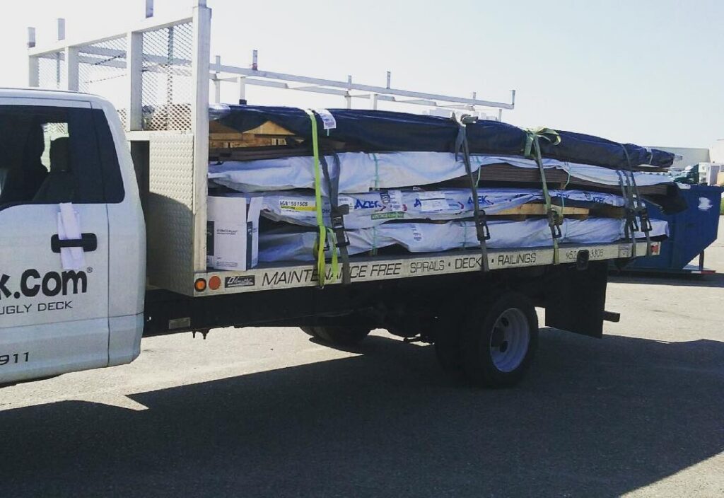 A flatbed truck loaded with large boxes and long DIY deck materials secured by straps, parked in a lot on a sunny day. Part of the truck’s cab, company branding for a deck builder, and a waste dumpster are visible in the background.