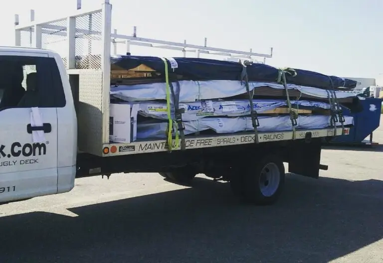 A flatbed truck loaded with large boxes and long DIY deck materials secured by straps, parked in a lot on a sunny day. Part of the truck’s cab, company branding for a deck builder, and a waste dumpster are visible in the background.