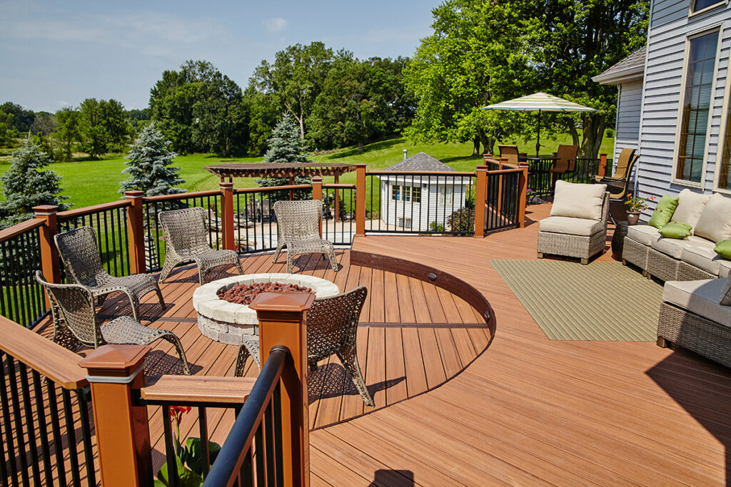 A spacious wooden deck—crafted by expert deck builders—features a circular fire pit, wicker chairs, an outdoor sofa with green cushions, a rug, and a table with an umbrella. The deck overlooks a green lawn bordered by trees.