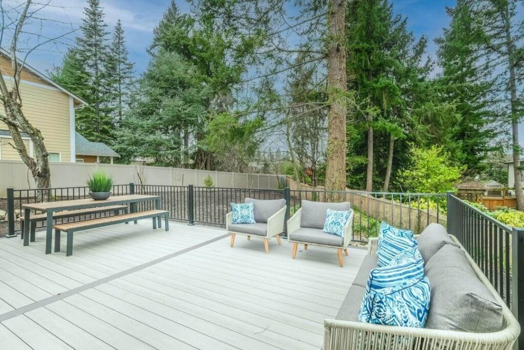 Spacious outdoor deck with gray flooring, modern gray patio furniture with blue patterned pillows, a wooden bench and table—expertly crafted by a professional deck builder—surrounded by trees and a tan fence.