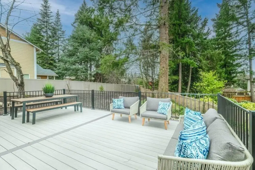 Spacious outdoor deck with gray flooring, modern gray patio furniture with blue patterned pillows, a wooden bench and table—expertly crafted by a professional deck builder—surrounded by trees and a tan fence.
