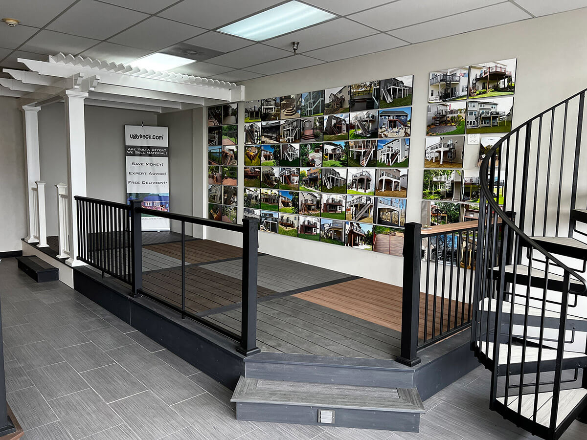 A showroom featuring a small deck display, railing, and pergola structure by a leading Deck Builder. Behind it, a wall showcases photos of outdoor living spaces. A spiral staircase is on the right, and the gray-tiled floor adds style.