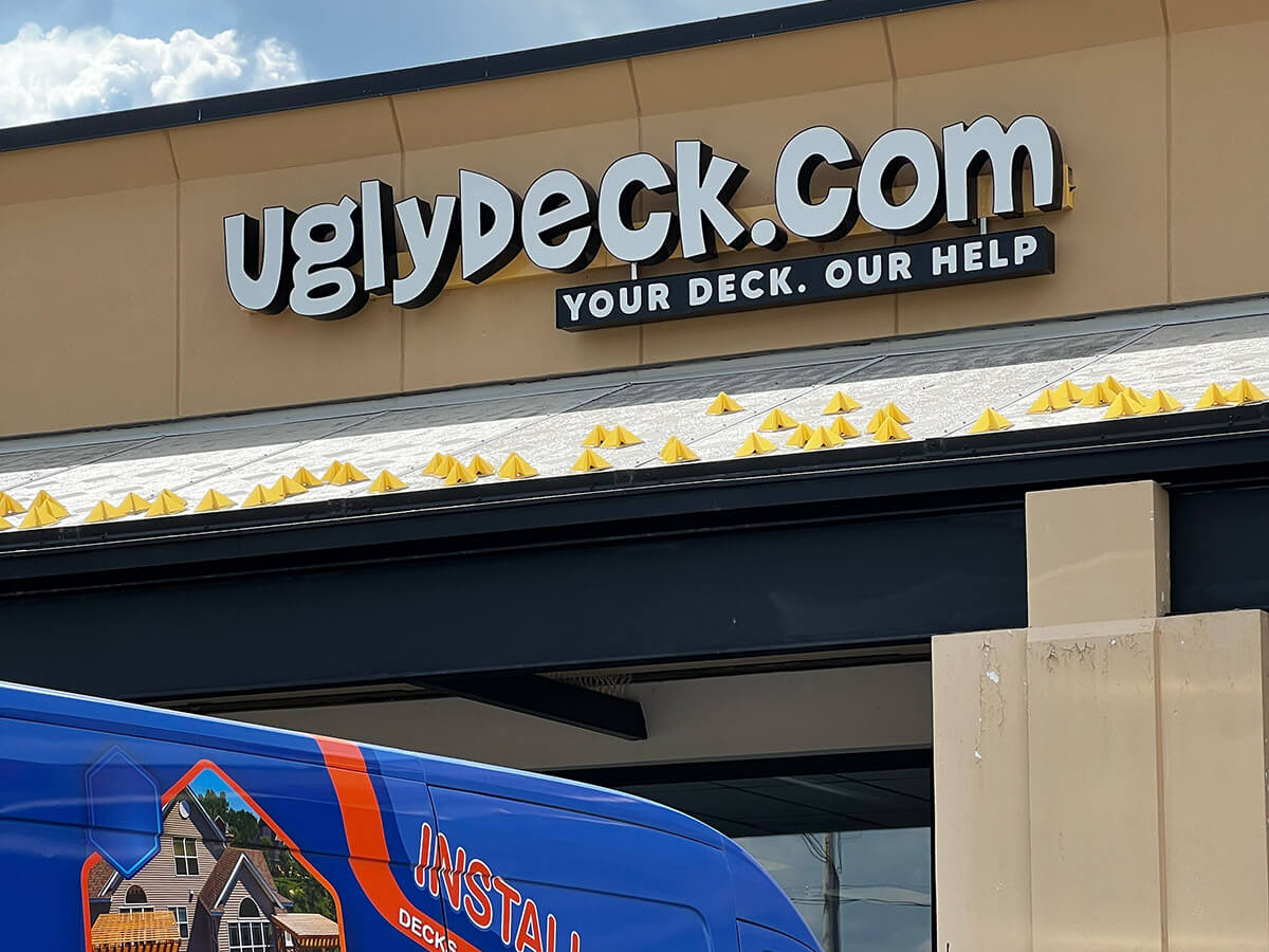 Storefront sign reads “UglyDeck.com Your Deck. Our Help” in bold letters; part of a blue and orange van with a house graphic is visible in front of the building. Yellow roof decorations stand out—perfect spot for DIY deck materials and expert deck builder help.