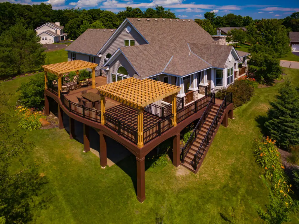Aerial view of a large house with a spacious elevated wooden deck, crafted with quality DIY deck materials, featuring two pergolas, outdoor seating areas, and a wide staircase, all surrounded by green grass, trees, and landscaping.