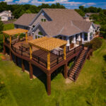 Aerial view of a large house with a spacious elevated wooden deck, crafted with quality DIY deck materials, featuring two pergolas, outdoor seating areas, and a wide staircase, all surrounded by green grass, trees, and landscaping.