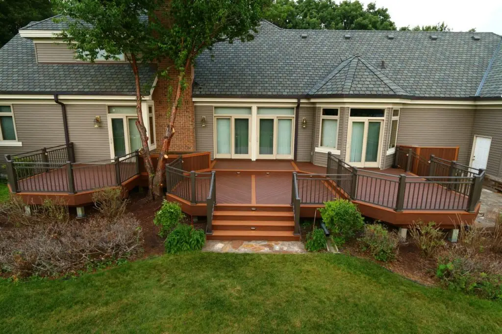 Spacious wooden backyard deck with steps, metal railings, and built-in lighting, attached to a modern house with large windows and gray siding—perfect for those seeking DIY deck materials or a professional deck builder.