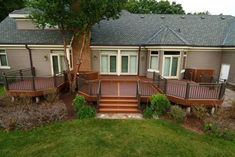 A spacious wooden backyard deck with railings, steps, and decorative landscaping showcases expert deck builder craftsmanship. Attached to a modern house with large windows and gray siding, a tree grows through the deck near the house.