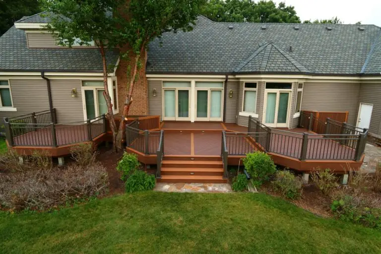 A spacious wooden backyard deck with railings, steps, and decorative landscaping showcases expert deck builder craftsmanship. Attached to a modern house with large windows and gray siding, a tree grows through the deck near the house.