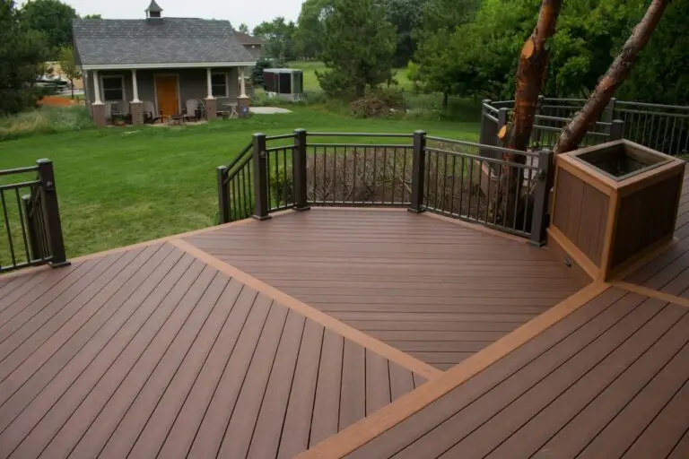 A spacious wooden deck with metal railings, built using quality DIY Deck Materials, overlooks a green backyard with a small house, trees, and lawn. The deck features a planter box around a tree and showcases the open, well-kept yard.