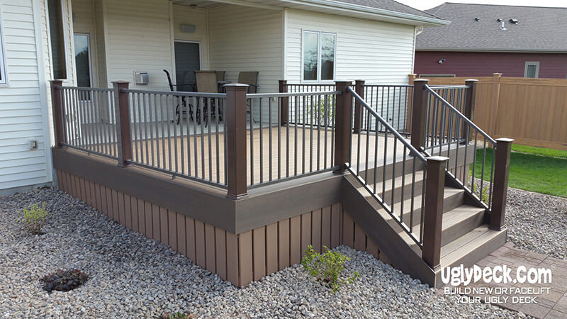 A raised backyard deck built with quality DIY Deck Materials features dark brown railing and steps, bordered by gravel landscaping and small shrubs, attached to a light-colored house with outdoor patio furniture visible.