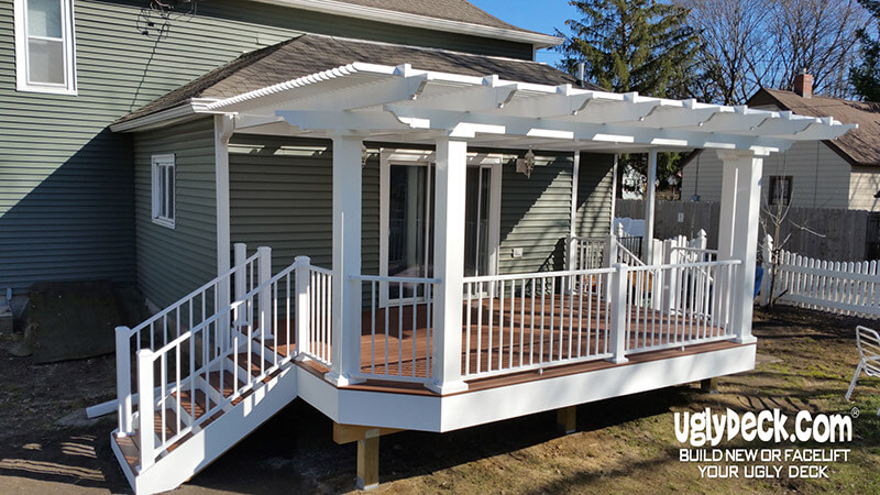 A modern white pergola with railings and stairs leads to a raised deck attached to a green house. This clean, well-maintained space showcases expert deck builder skills, with the uglydeck.com logo in the corner.