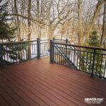 A brown wooden deck with black railings, built using quality DIY Deck Materials, overlooks a snowy, wooded backyard filled with trees and evergreens. Stairs lead down from the deck. The uglydeck.com logo appears in the bottom right corner.