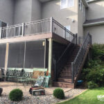 A two-story house with a raised deck built by a skilled deck builder features stairs leading to a stone patio. The patio offers multiple chairs and tables, while shrubs and rocks border the area under the screened-in deck.