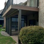 A modern house with a raised deck install featuring black railings and a covered patio area underneath. The landscaping includes bushes and decorative rocks along the edge, showcasing the work of an expert deck builder.