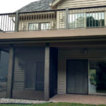 A beige two-story house features a screened-in porch below and an upper balcony with dark railings—a perfect showcase for quality DIY deck materials. Landscaping with rocks and shrubs surrounds the inviting porch area.