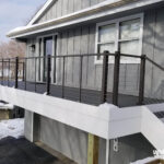 A modern home deck with dark railing and cable wires extends from a gray house. Expert deck builder craftsmanship is evident in the elevated design, white trim, double windows, and stairs on the right, while snow covers the deck and ground.