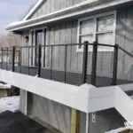 A modern home deck with dark railing and cable wires extends from a gray house. Expert deck builder craftsmanship is evident in the elevated design, white trim, double windows, and stairs on the right, while snow covers the deck and ground.