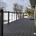 A modern gray composite deck, built with premium DIY deck materials and cable railing, overlooks a snowy landscape and frozen lake. Bare trees surround the space, and the deck attaches to a light-sided house. UglyDeck.com is branded in the lower right corner.