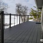 A modern gray composite deck, built with premium DIY deck materials and cable railing, overlooks a snowy landscape and frozen lake. Bare trees surround the space, and the deck attaches to a light-sided house. UglyDeck.com is branded in the lower right corner.