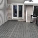 A gray composite deck with black railing, crafted with quality DIY Deck Materials, is attached to a house with beige siding. Two sliding glass doors lead outside, and some snow rests on the roof. The deck is empty.